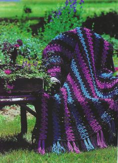 a purple and blue crocheted blanket sitting on top of a wooden bench next to flowers