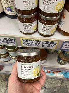 a hand holding up a jar of green tea spread in front of shelves with other jars