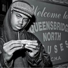 a black and white photo of a man holding a hot dog in front of a welcome to queensbridge north sign