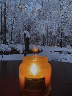 a candle sitting on top of a table in front of snow covered trees and bushes