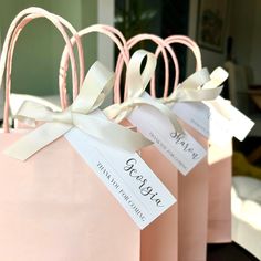 three pink paper bags with white ribbons and name tags on them, sitting on a table