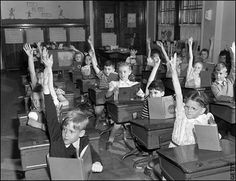 black and white photograph of children in classroom raising their hands up to the teacher's desk