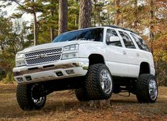 a white truck parked in the woods with large tires on it's front wheels