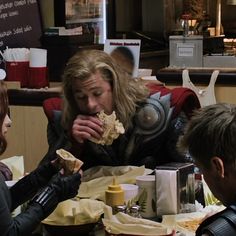 some people sitting at a table eating food and talking to one person dressed as thor