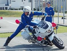 a man in blue jumpsuits leaning on a white motorcycle while another man stands behind him