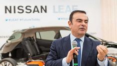 a man speaking into a microphone in front of a nissan leaf sign and an electric car