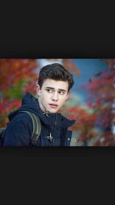 a young man is standing in front of a tree with red leaves and wearing a black jacket