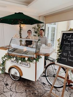 a man and woman behind a food cart with an umbrella over it that is selling pastries