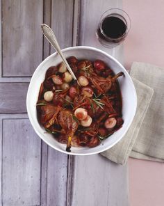 a white bowl filled with meat and vegetables next to a glass of wine on top of a table