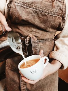 a person pouring coffee into a white cup with the word w on it and a spoon in their hand