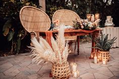 an outdoor table with candles, flowers and plants in baskets on the ground next to two wicker chairs