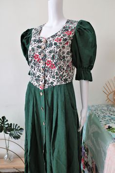 Stunning True Vintage Austrian trachten midi dress from the 1990s. Comfortable cotton linen blend fabric. Dark green sleeves and skirt, floral top. Fitted top, wide skirt. Sweetheart neckline, short puff sleeves with elasticated cuffs. Lined torso, button front, wooden trachten buttons, two side pockets. Made in Austria! BRAND: Wenger ERA: 1990s COLOR: Dark green, off-white, red, black Fabric: 60% cotton, 20% linen, 20% modal; lining 100% cotton SIZE: No size tag anymore, fits best a modern size Summer Cotton Prairie Dress With Gathered Sleeves, Cotton Midi Length Prairie Dress For Garden Party, Cotton Floral Print Prairie Dress With Short Sleeves, Summer Cotton Prairie Dress With Puff Sleeves, Green Puff Sleeve Dress In Cottagecore Style, Folk Style Green Embroidered Dress With Floral Details, Green A-line Cotton Vintage Dress, Cotton Prairie Dress For Garden Party, Midi Length, Vintage Austrian Dress