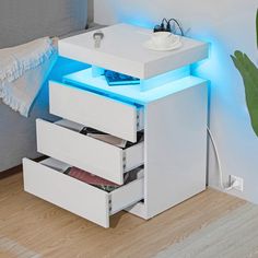 a white night stand with blue lights on top and drawers underneath the table, next to a plant