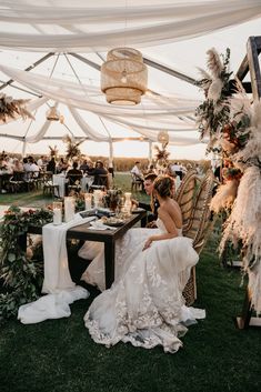 a woman sitting at a table in front of a white tent with flowers and greenery