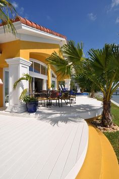 a patio with chairs and palm trees next to the ocean in front of a yellow house