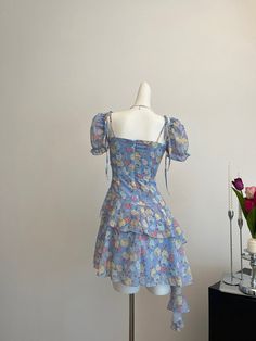 Circuitous and crafted with care, this blue floral dress weaves its way through timeless fashion with a modern twist. The layers of lightweight fabric create a gentle cascade, giving this dress a buoyant, ethereal quality that’s perfect for spring and summer events. Decorated with a vibrant array of floral patterns in soft hues, the dress features dainty shoulder straps and a rosette detail for a touch of romance. Its asymmetrical hemline and flowing silhouette ensure it moves gracefully with th Summer Tiered Dress For Garden Party, Light Blue Mini Dress With Ruffles And Square Neck, Light Blue Square Neck Mini Dress With Ruffles, Fitted Floral Print Chiffon Dress For Summer, Light Blue Fitted Chiffon Dress For Summer, Fitted Chiffon Summer Dress For Spring, Spring Floral Print Mini Chiffon Dress, Blue Summer Mini Dress For Garden Party, Blue Chiffon Mini Dress For Summer