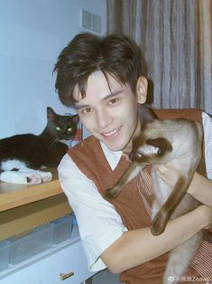 a young man holding a siamese cat in front of a black and white cat