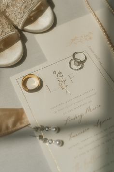 two wedding rings on top of a white paper with gold trimmings next to a pair of bride's shoes