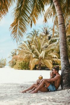 a man and woman sitting under a palm tree on the beach with drinks in their hands