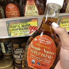 a person holding up a bottle of maple syrup in front of some other bottles on the shelf