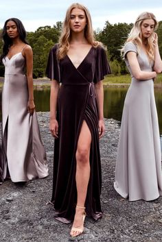 three women in long dresses standing next to each other on rocks near water and trees