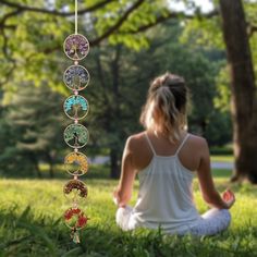 a woman sitting on the grass in front of a wind chime with seven chakras hanging from it