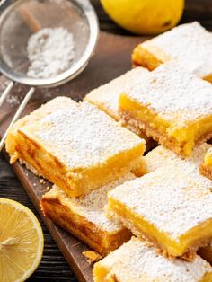 lemon bars with powdered sugar on a cutting board next to sliced lemons and a glass of water