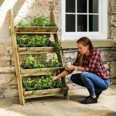 a woman kneeling down next to a wooden planter filled with green plants and herbs