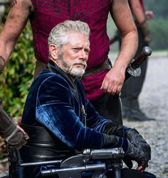 an older man sitting in a wheel chair with other people around him and holding onto the handlebars