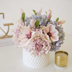 a white vase filled with pink and purple flowers next to a gold candle on a table