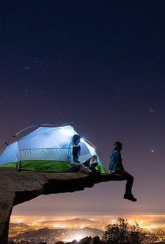 two people sitting on top of a cliff next to a tent at night with stars in the sky