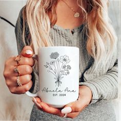 a woman holding a white coffee mug with flowers on the front and bottom in her hands