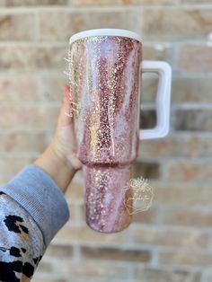 a person holding up a pink and gold glittered coffee cup in front of a brick wall