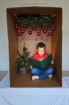 a young boy is sitting in a cardboard box reading a book while surrounded by christmas decorations