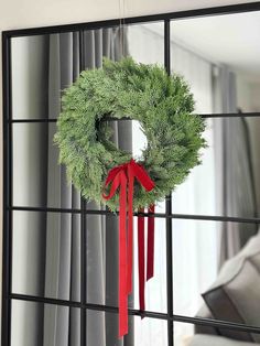 a christmas wreath hanging on a mirror in front of a window with red ribbon tied around it