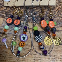 A Halloween, spooky earring set! This set of 3 earrings is a handmade product. It includes 3 dangle earrings of varying haunted designs.  1. The first pair, has purple, green, and orange beads. The beads say "Boo" on an orange background. It has two different colored dangle charms at the end. One is a green lollipop and one is a purple lollipop. The metal for this pair is gold and bronze. It features a metal, wire earring hook.  2. The second pair are tear dropped shaped. There is a silver, metal tear drop shaped outer section with a line of beads in the middle on a wire. The beads are purple, orange, green, black, and metallics. There are  spider, heart, and checkered black and white heart beads. The metal for this pair is silver. It features a metal, small hoop earring hook that clasps i Beaded Dangle Earrings For Halloween, Dangle Beaded Earrings For Halloween, Handmade Dangle Earrings For Halloween, Fun Dangling Beads Jewelry For Gifts, Fun Dangling Beads Jewelry As Gift, Fun Jewelry With Dangling Beads For Gift, Halloween Beaded Dangle Earrings Gift, Halloween Gift Beaded Dangle Earrings, Handmade Dangle Halloween Jewelry