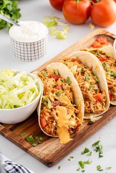 three tacos with shredded chicken, lettuce and tomatoes on a cutting board