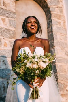 a woman standing in front of a stone building holding a bouquet of flowers and smiling