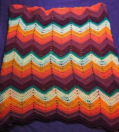 a crocheted blanket is laying on a purple surface with an orange and green zigzag pattern