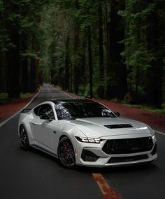 a white sports car driving down the road in front of some tall trees and greenery