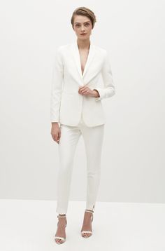 a woman in a white suit and heels standing on a white surface with her hands behind her back