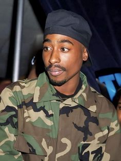 a man in camouflage jacket and hat looking at the camera with people watching from behind him