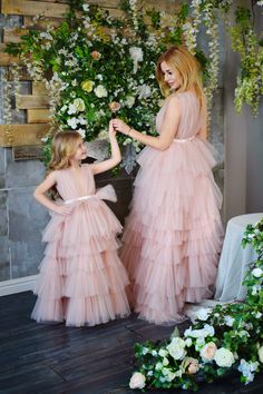 two girls in pink dresses standing next to each other with flowers on the wall behind them