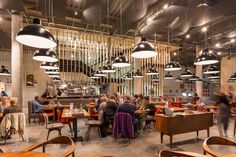 people sitting at tables in a restaurant with lights hanging from the ceiling and wooden chairs