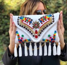 a woman holding up a white clutch bag with tassels and beads on it