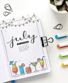 a notebook with the word july written on it next to markers, pens and potted plants