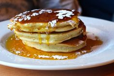 stack of pancakes covered in syrup and powdered sugar on a white plate sitting on a wooden table