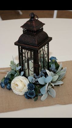 a lantern with flowers and greenery on top of a cloth table runner at a wedding reception