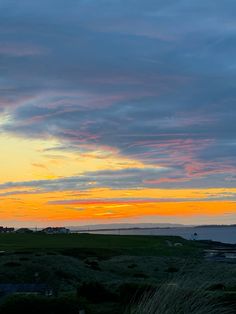 the sun is setting over an ocean with grass and houses in the foreground, as seen from across the water