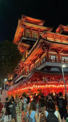 many people are walking around in front of a tall building at night with red lights on it